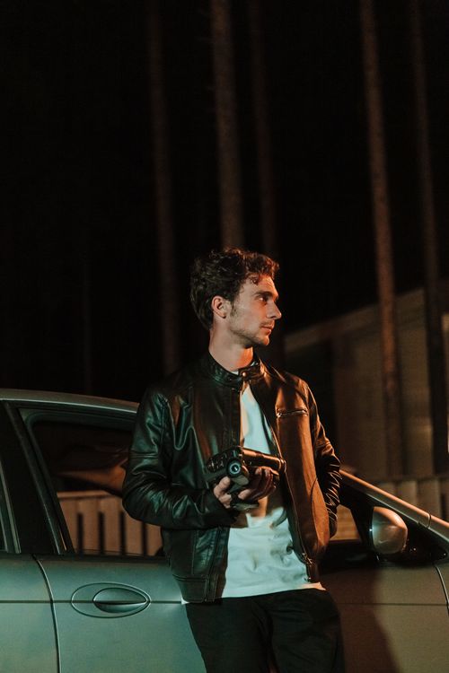 Free A man in a leather jacket leans on a car while holding a camcorder during nighttime. Stock Photo