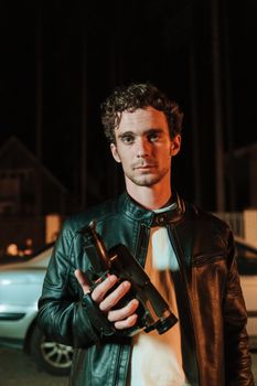 Young man in a leather jacket holding a camcorder at night with a car in the background.