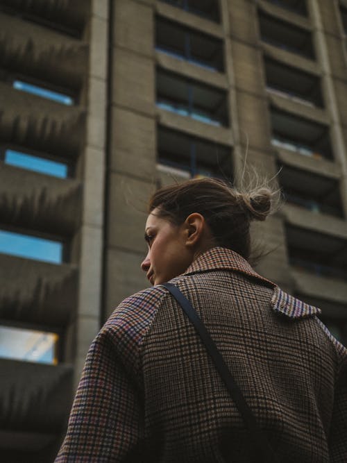 Free Woman with Tied Hair in Checkered Coat Standing Before Tall Building Stock Photo