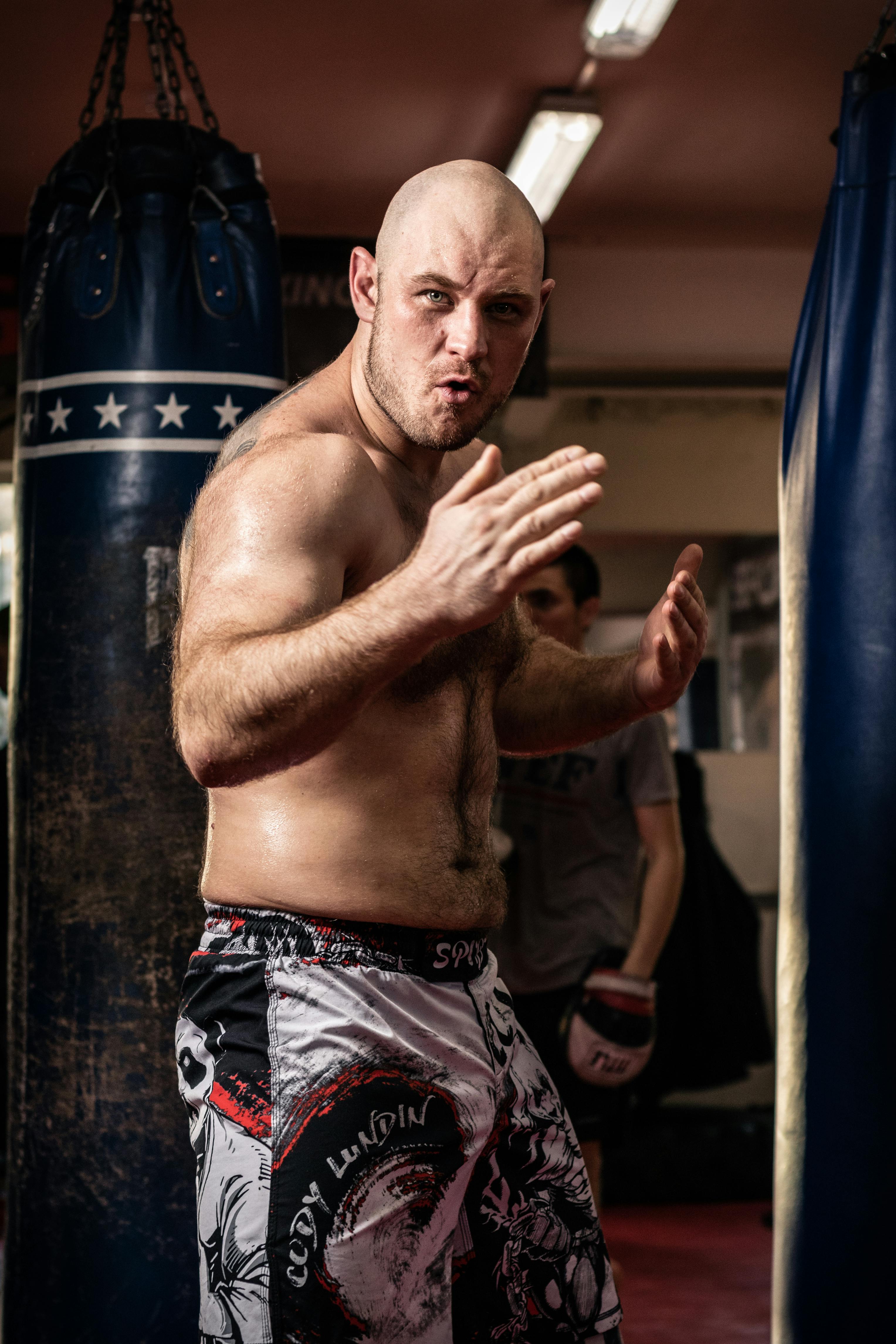 Shirtless man engages in martial arts training with a punching bag in a gym setting.