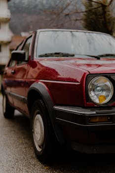 Close-up of a vintage red car parked outdoors on a rainy day with a blurred background.