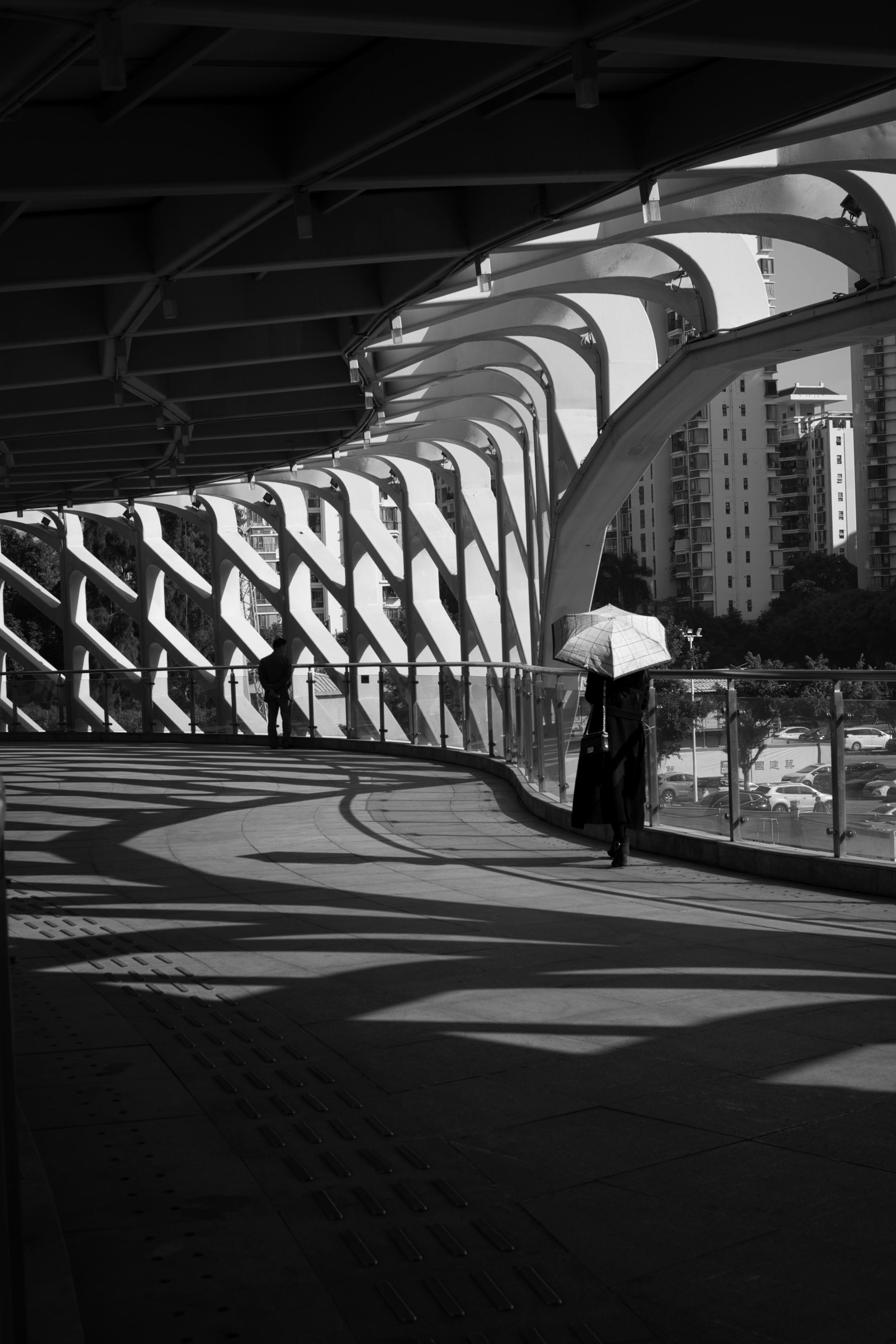 Elegant black and white image of a futuristic bridge pathway with shadows and an umbrella-carrying pedestrian.