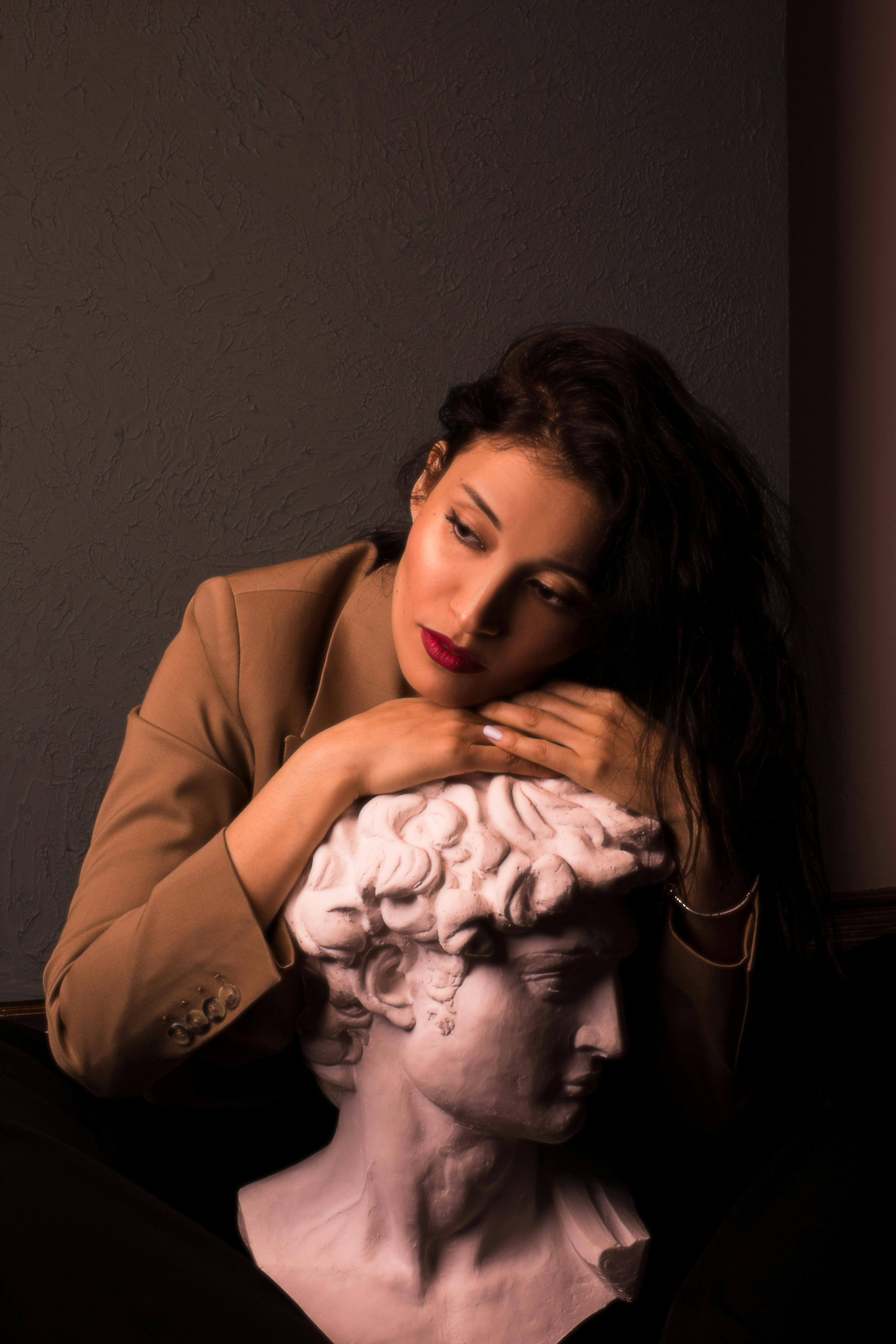 A contemplative woman in a brown blazer rests her head on a classical sculpture bust.