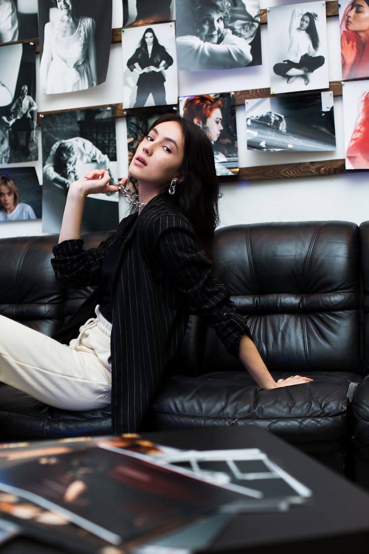 Brunette Woman Sitting On Black Leather Sofa In Photo Studio And Playing With Necklace