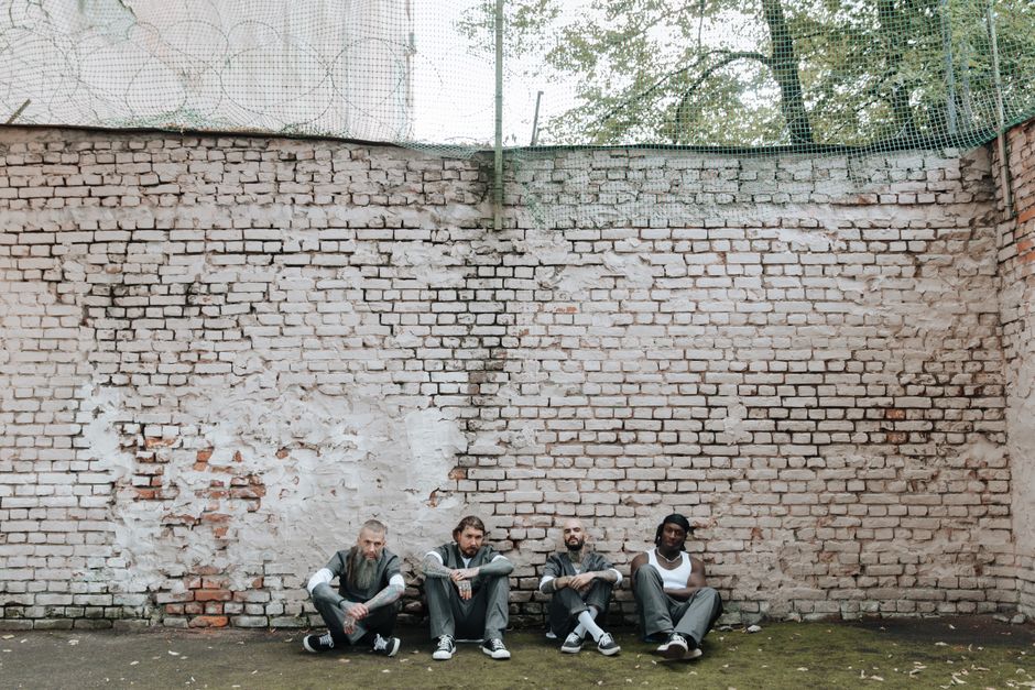 Four men sitting casually against a brick wall outdoors, showcasing diversity and urban style. Four men sitting casually against a brick wall outdoors, showcasing diversity and urban style.