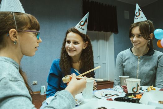 Teenagers celebrate a birthday indoors with party hats and food, sharing smiles and conversation.