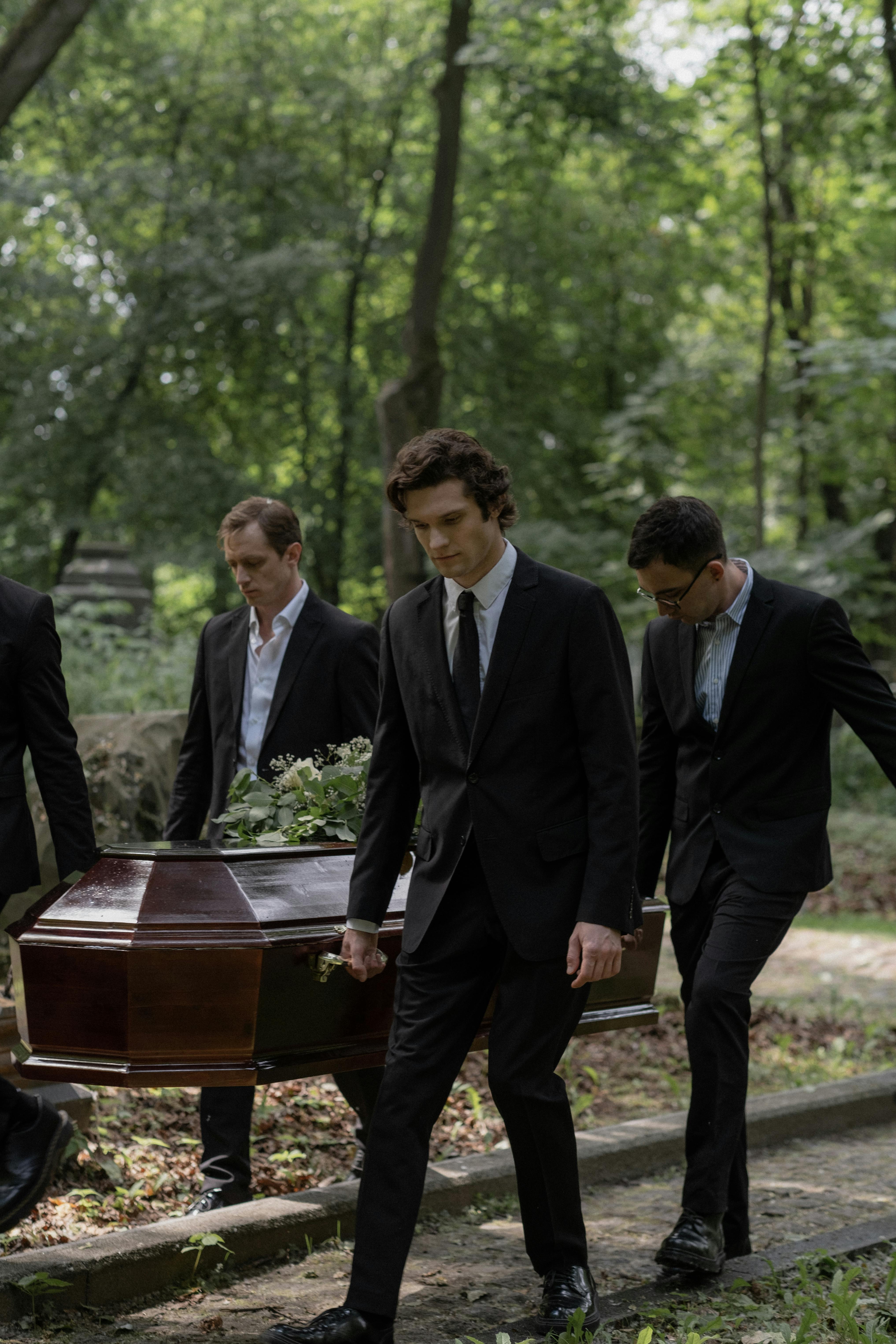 Men in suits solemnly carry a coffin through a peaceful forest cemetery during a daytime funeral procession.