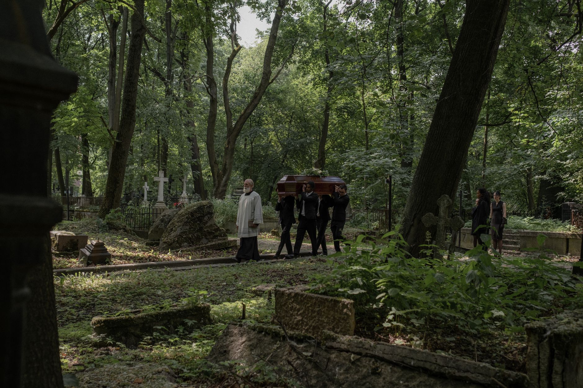 A group carries a coffin through a peaceful, wooded cemetery during a solemn funeral procession.