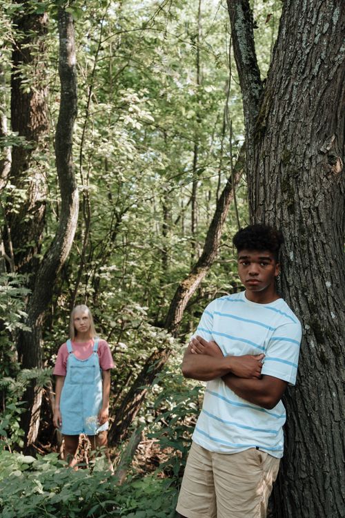 Free Two teenagers in a forest, one leaning against a tree, representing friendship and adventure. Stock Photo