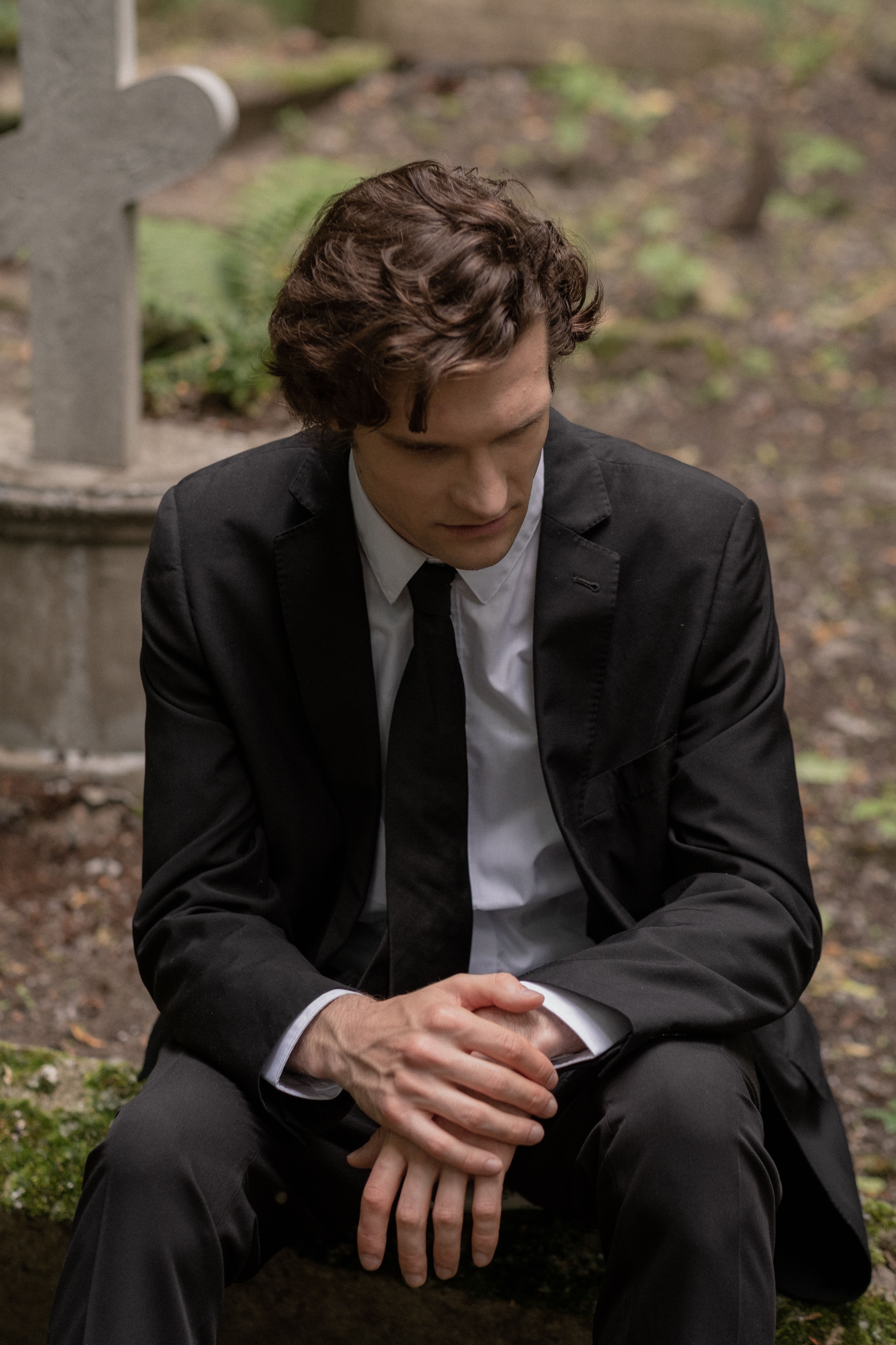 A man in a black suit sits pensively in a cemetery, evoking themes of loss and reflection.