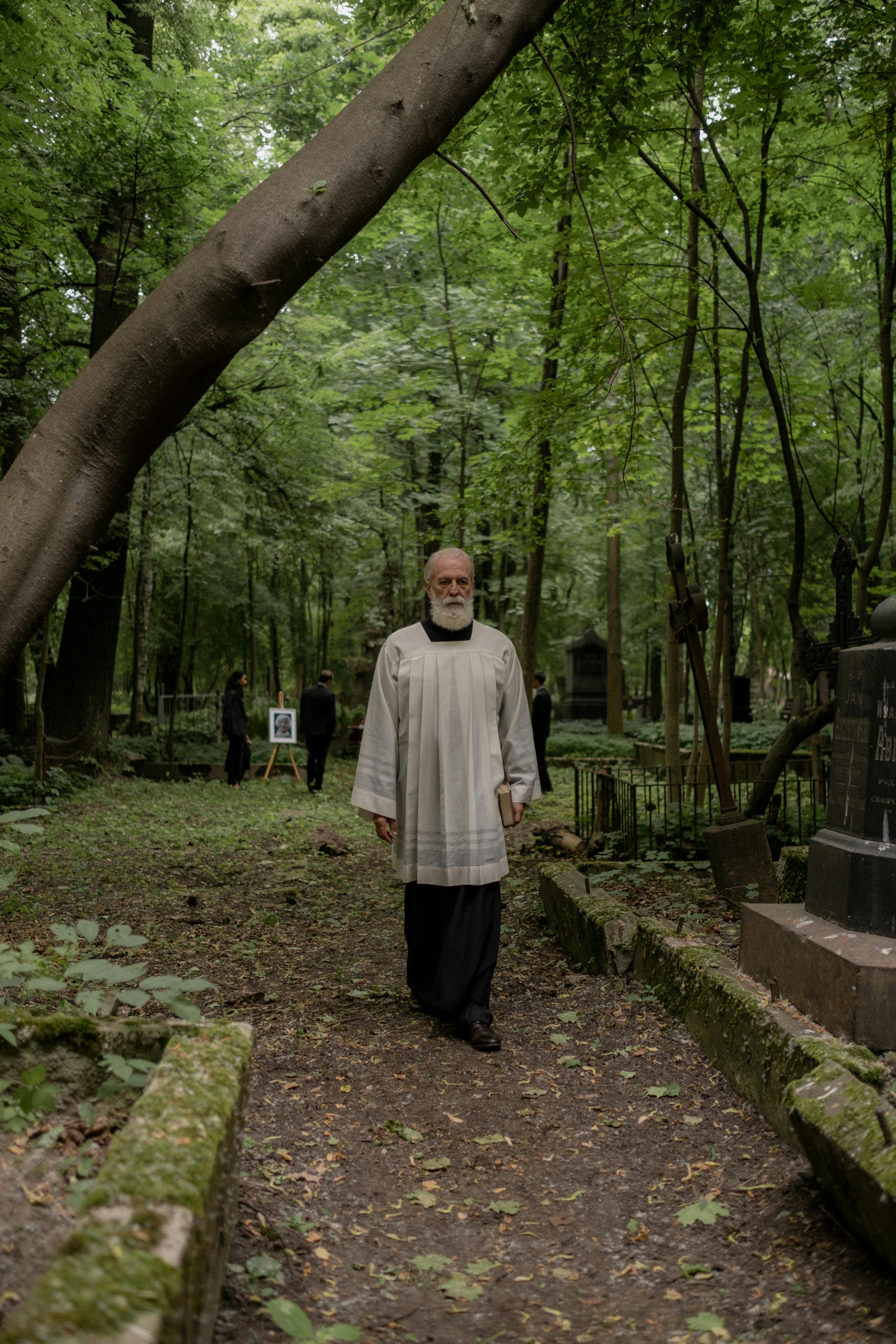 A bearded pastor in robes walks through a tranquil forest cemetery, surrounded by lush greens.
