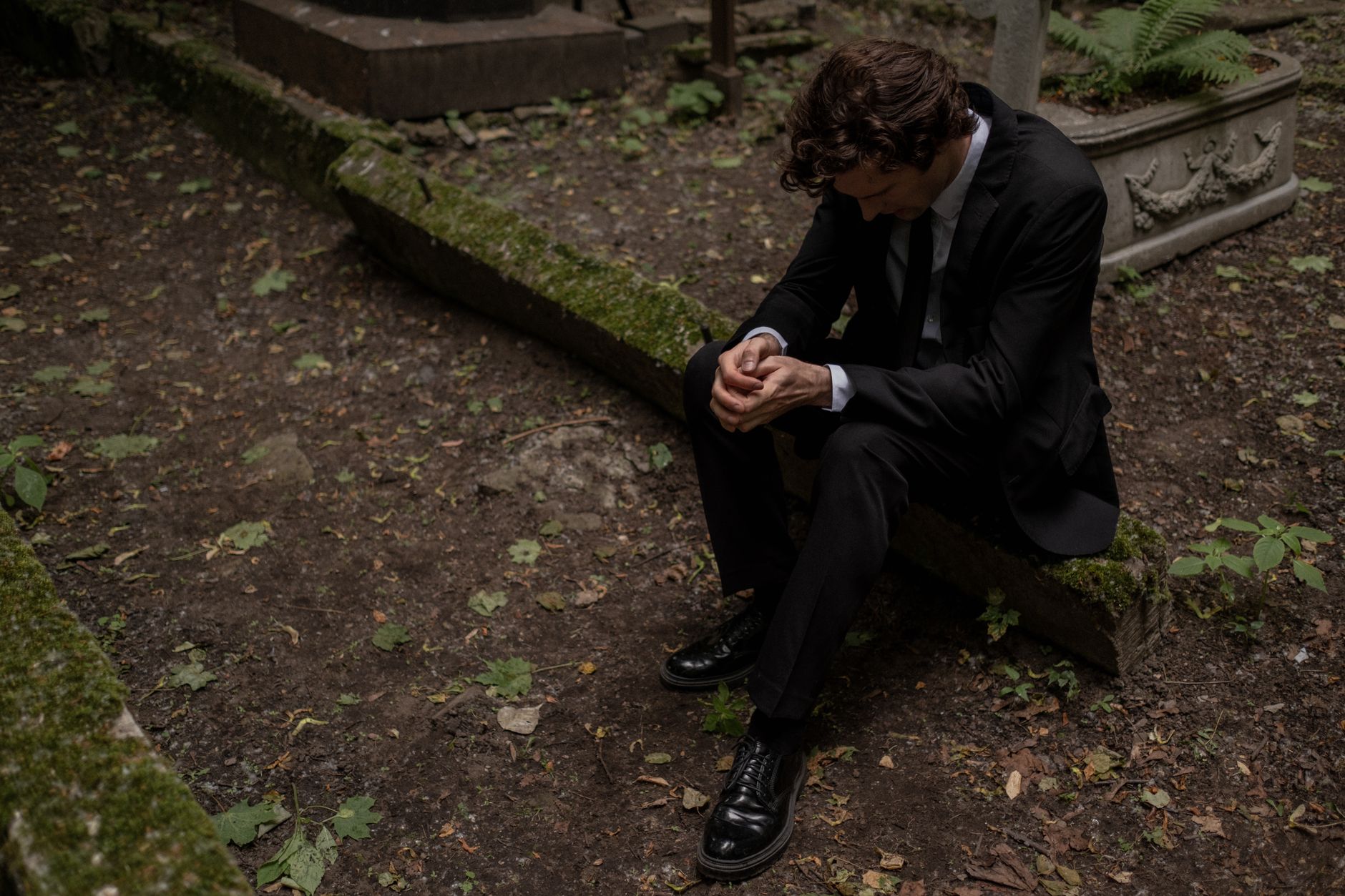 A man in a black suit sitting in a cemetery, reflecting loss and mourning.