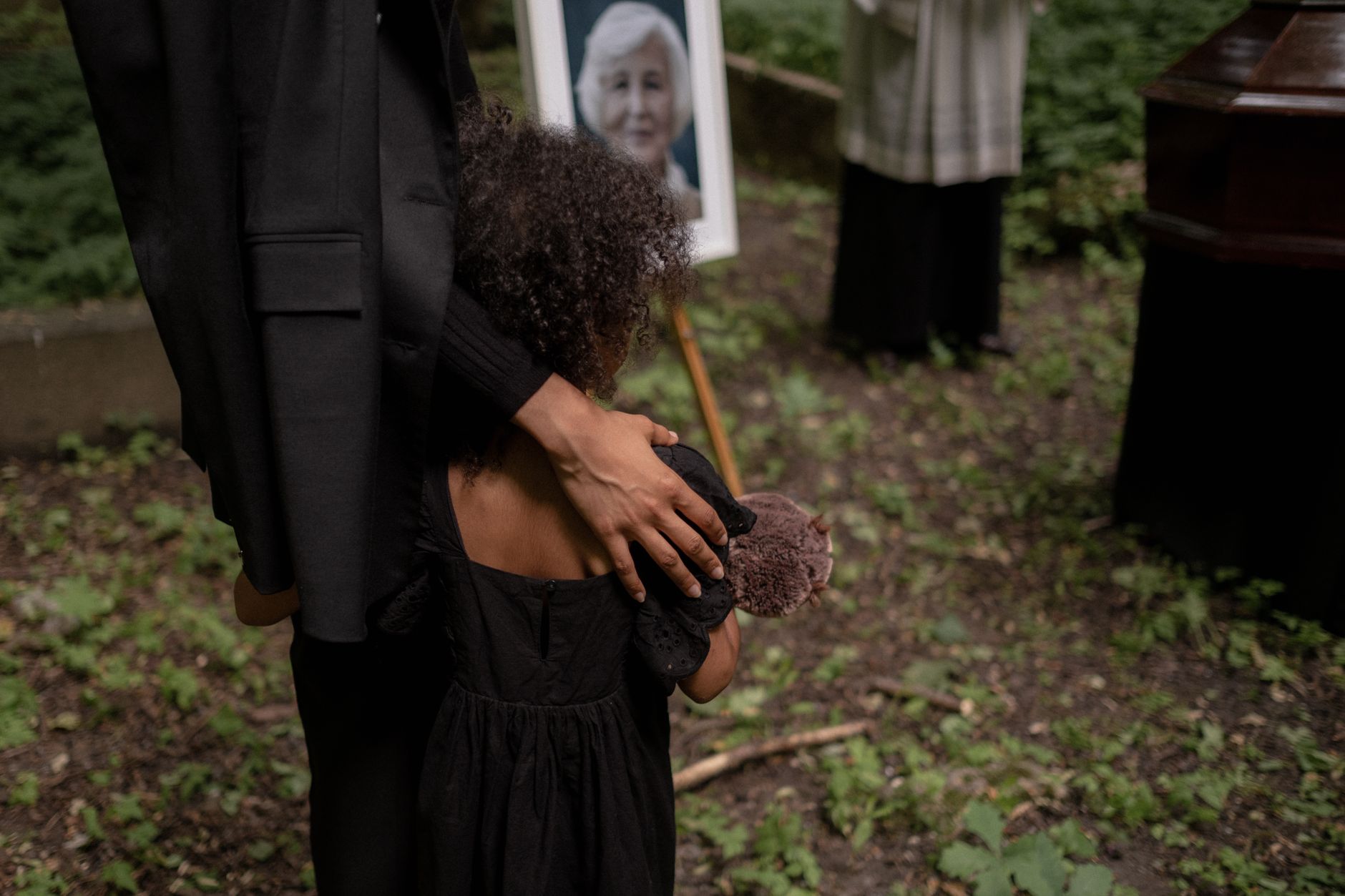 A child in black dress holds a toy bear, attending a funeral ceremony in a cemetery.