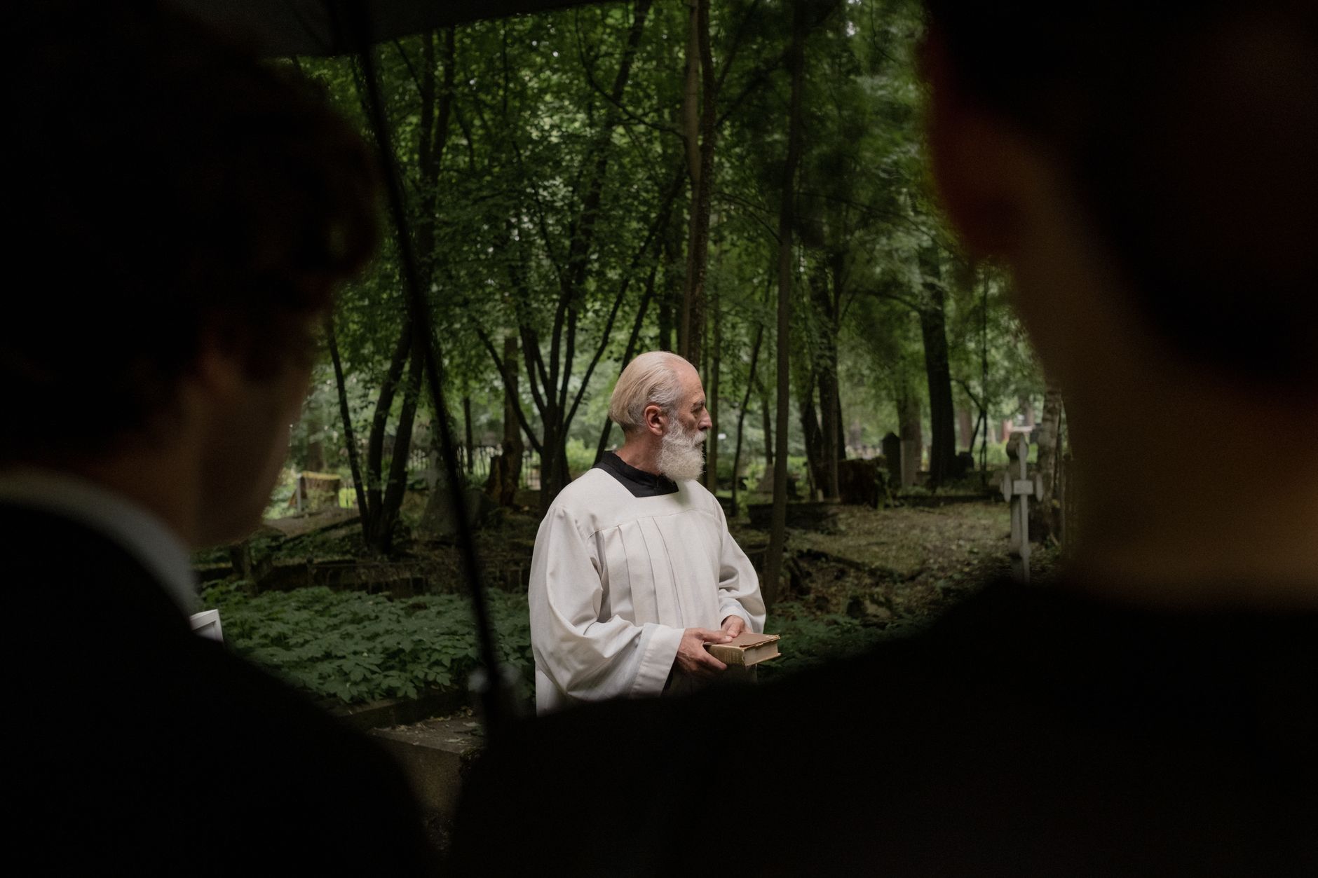 An elderly priest conducts a funeral in a tranquil forest cemetery.