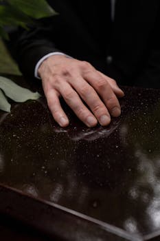 A man's hand rests on a wet wooden table beside green leaves. Artistic and moody.