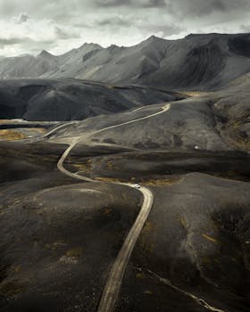 A solitary winding road snakes through a dramatic, rugged mountainscape under a cloudy sky.