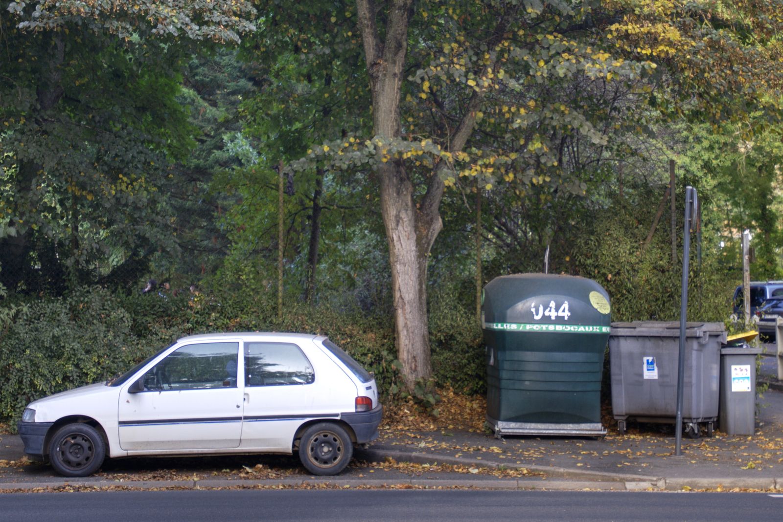 Voiture blanche garée près de conteneurs de recyclage sous des arbres d'automne