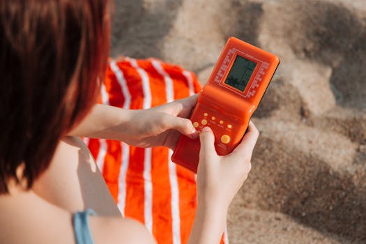 A person playing a vintage orange handheld game console on a sandy beach.