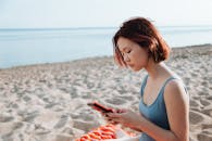 穿着泳衣的年轻女子坐在海滩岸边使用智能手机