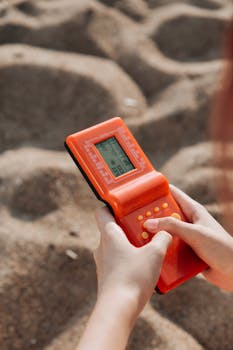 Hands holding an orange game console in a sandy environment, close-up view.