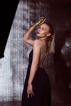 A stylish woman in a backless black dress poses in a studio with golden makeup accents.