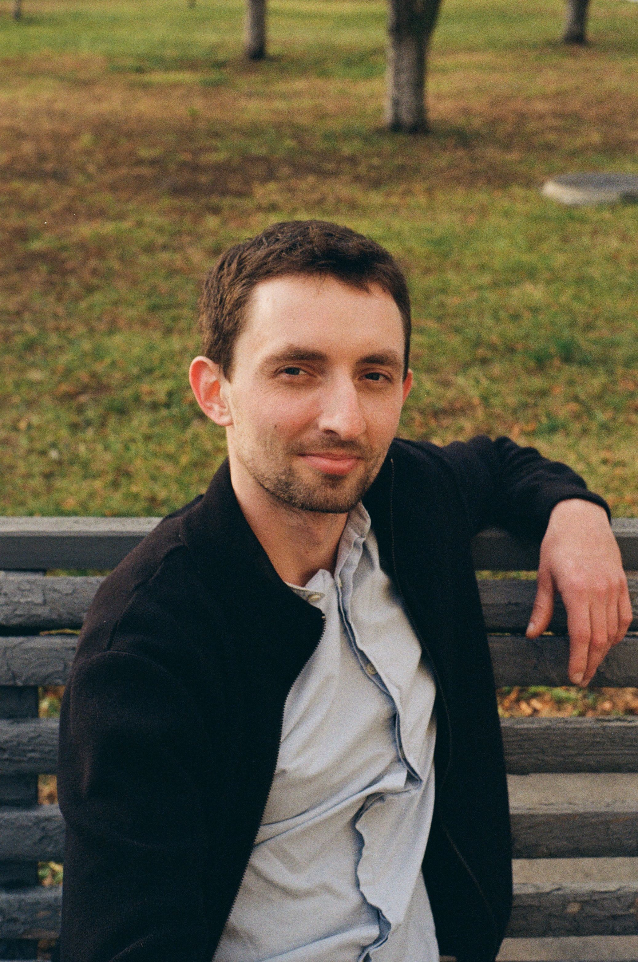 Man in black jacket sitting on bench in outdoor park setting.