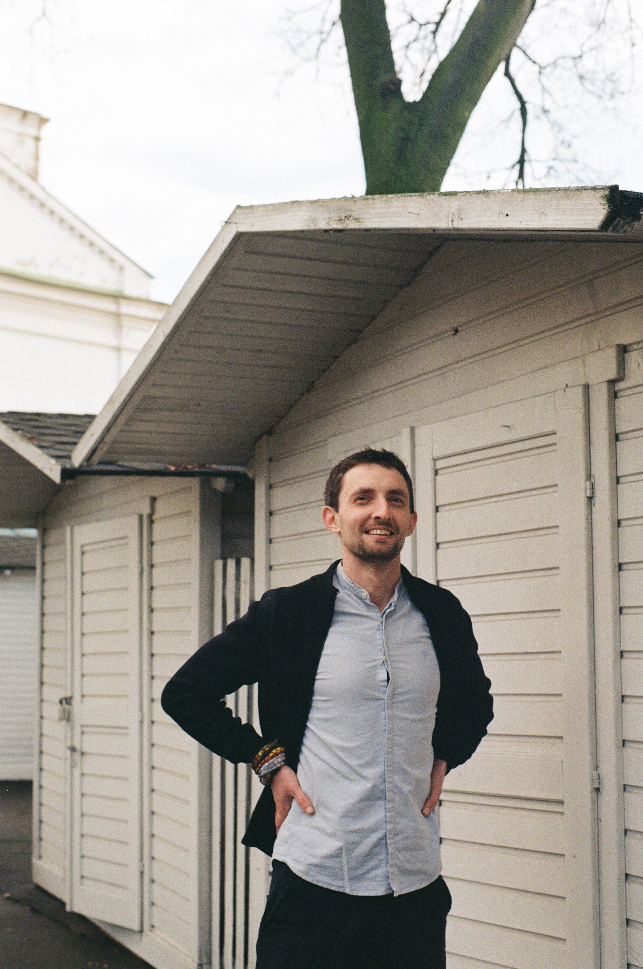 A smiling man in a black jacket stands outdoors with hands on waist, exuding confidence.