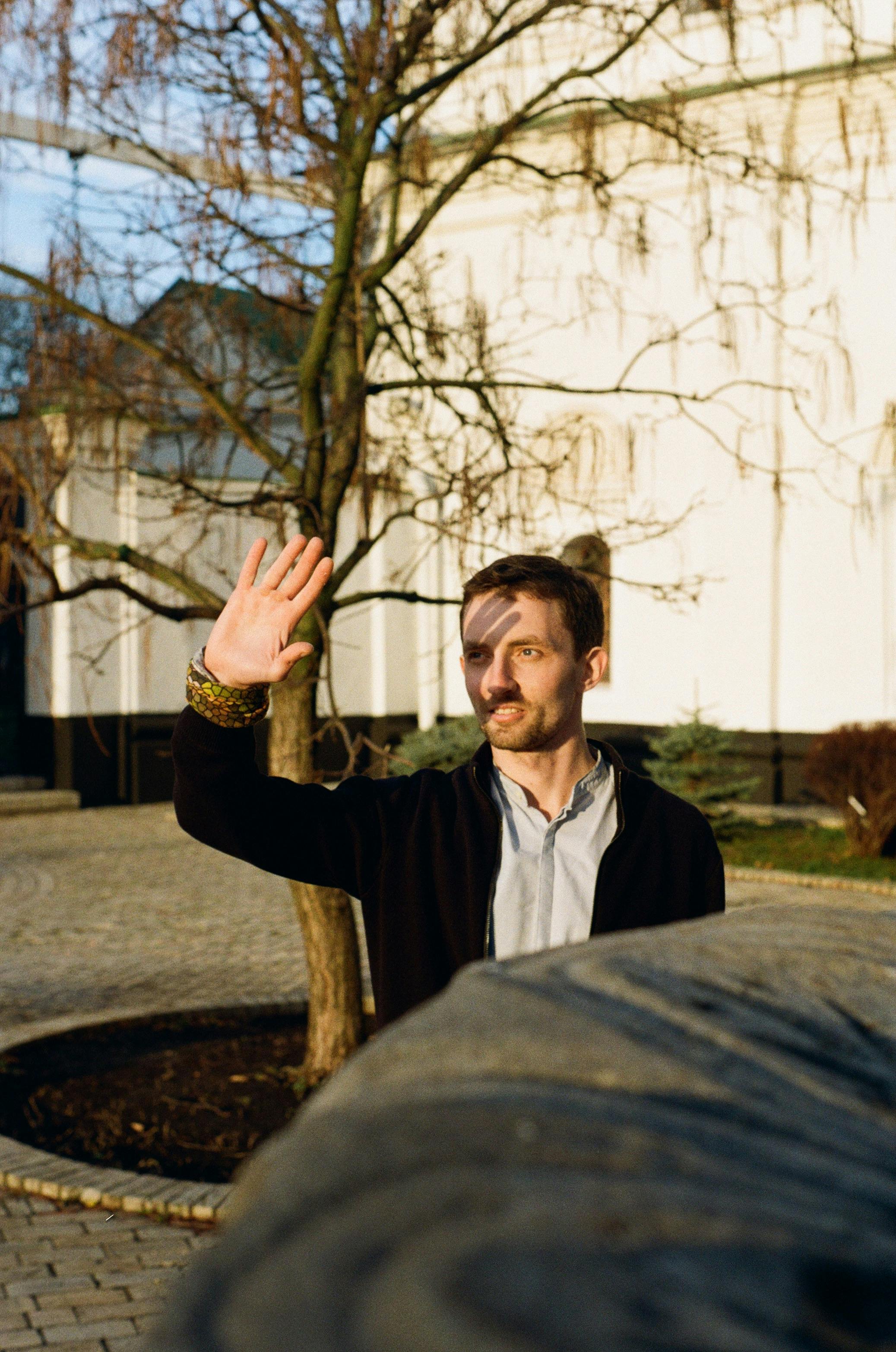 A man in a black jacket stands outside during the day, raising his hand in the sunlight against a leafless tree backdrop.