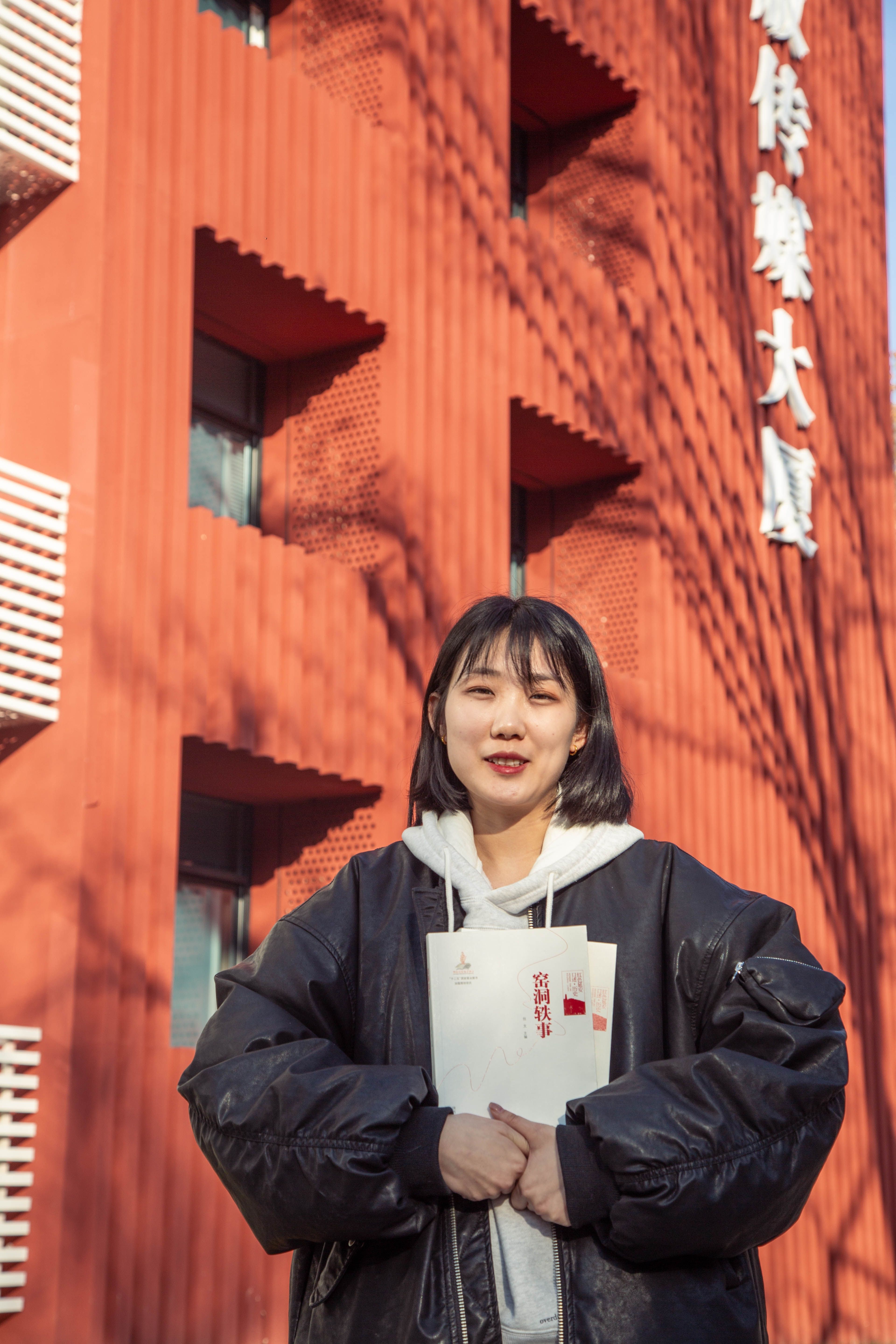 Smiling young woman in a black jacket holding books against a modern urban building backdrop.