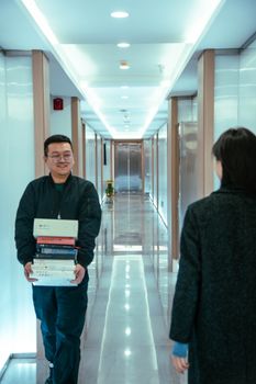 A smiling man walks in a sleek office corridor, carrying a stack of books and interacting with a woman.