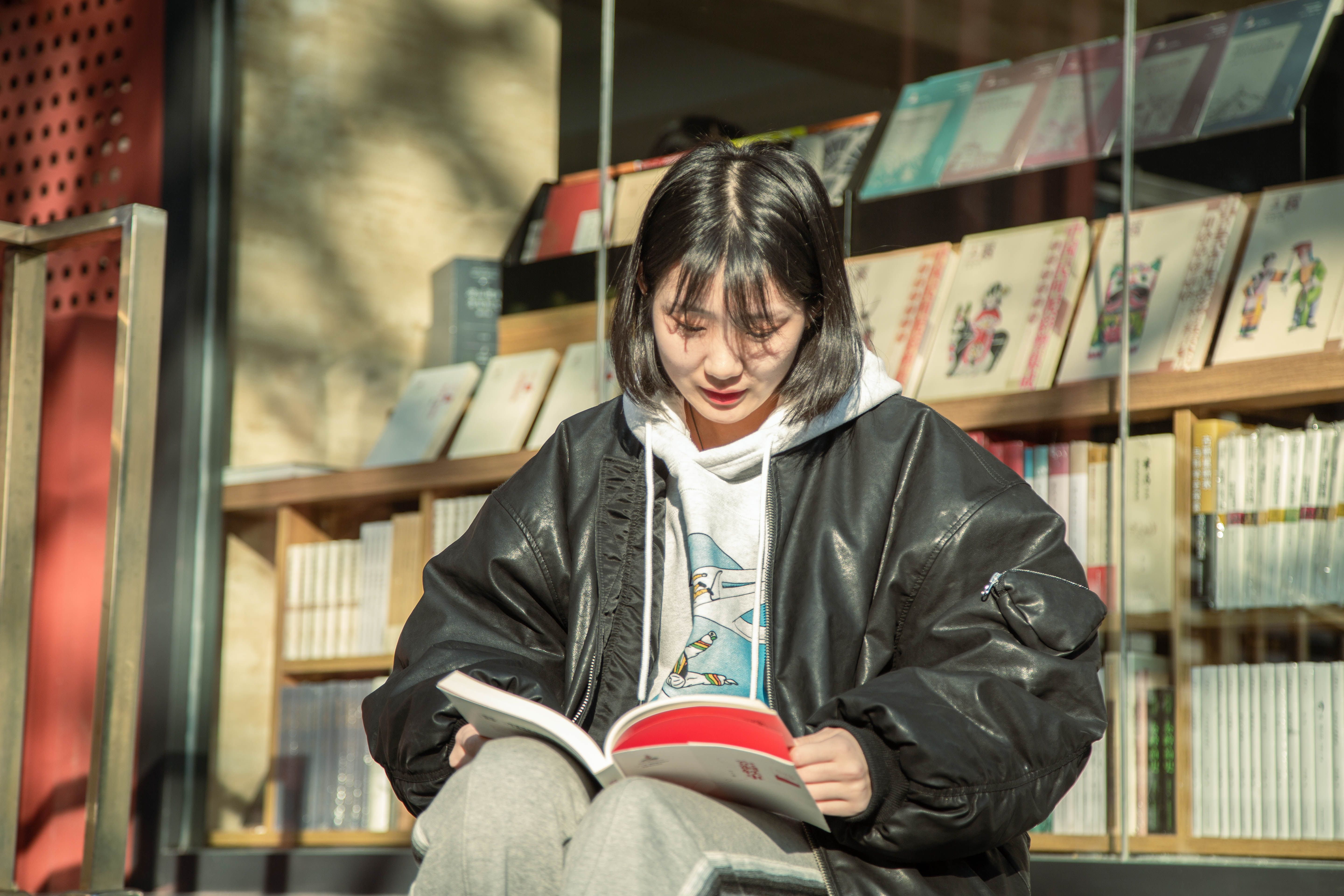 Asian woman reading outside a library, wearing a black jacket, in sunlight.