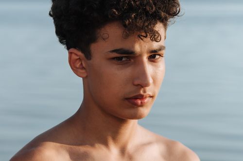 Free Portrait of a young man with curly hair standing shirtless by the lake. Stock Photo