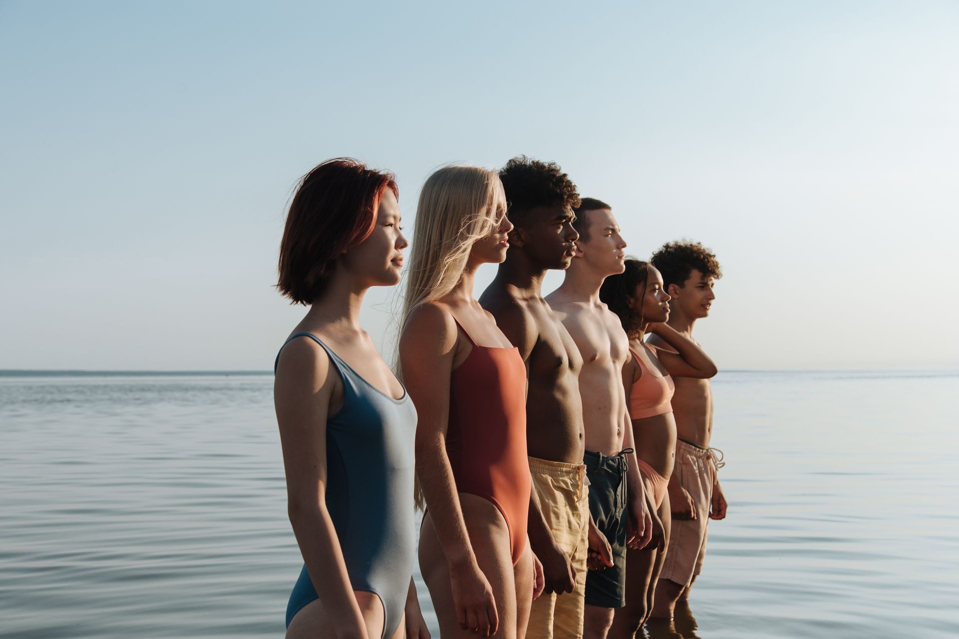 A diverse group of teenagers standing side by side in swimsuits by the seaside during summer.