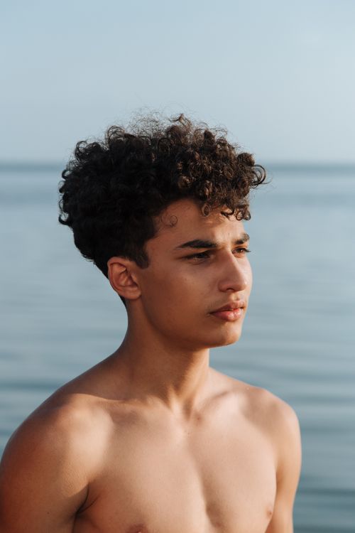 Free Portrait of a shirtless teen with curly hair standing by the sea. Summer and carefree vibes. Stock Photo