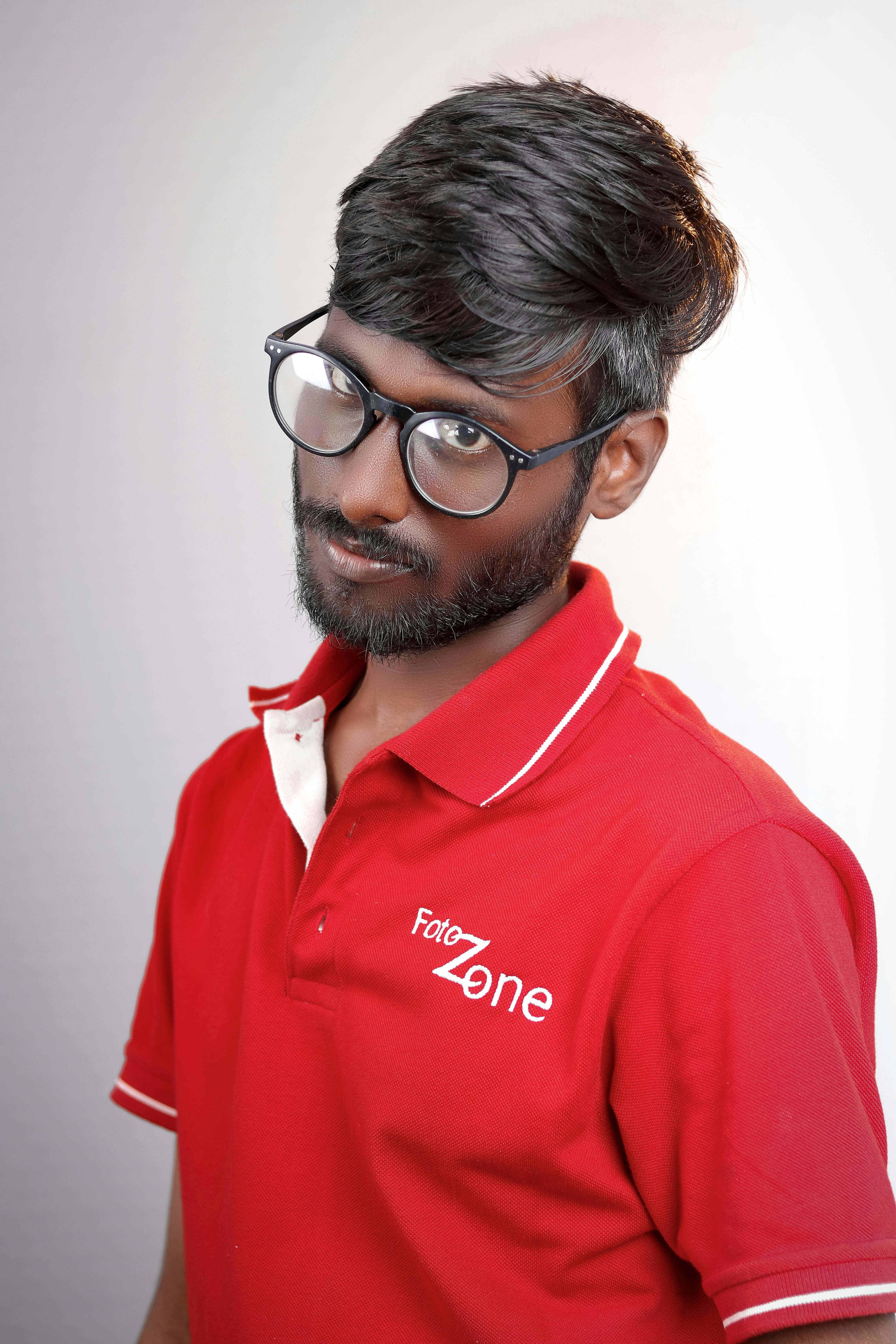 A confident South Asian man with eyeglasses in a red polo shirt posing against a neutral background.