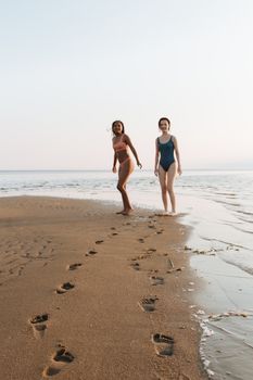 Two women in swimwear walking along the sandy beach during summer, creating footprints in the sand.