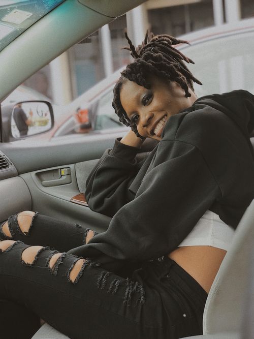 Free Happy woman with dreadlocks smiling inside a car, wearing a black hoodie and ripped jeans. Stock Photo