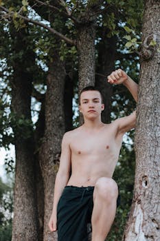 Shirtless teenager leaning on a tree trunk during summer, enjoying nature.