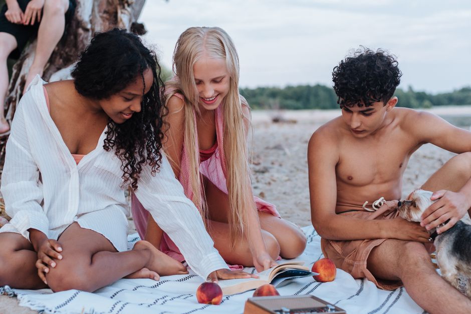 A diverse group of friends on a beach picnic, reading and enjoying fruit with a small dog.