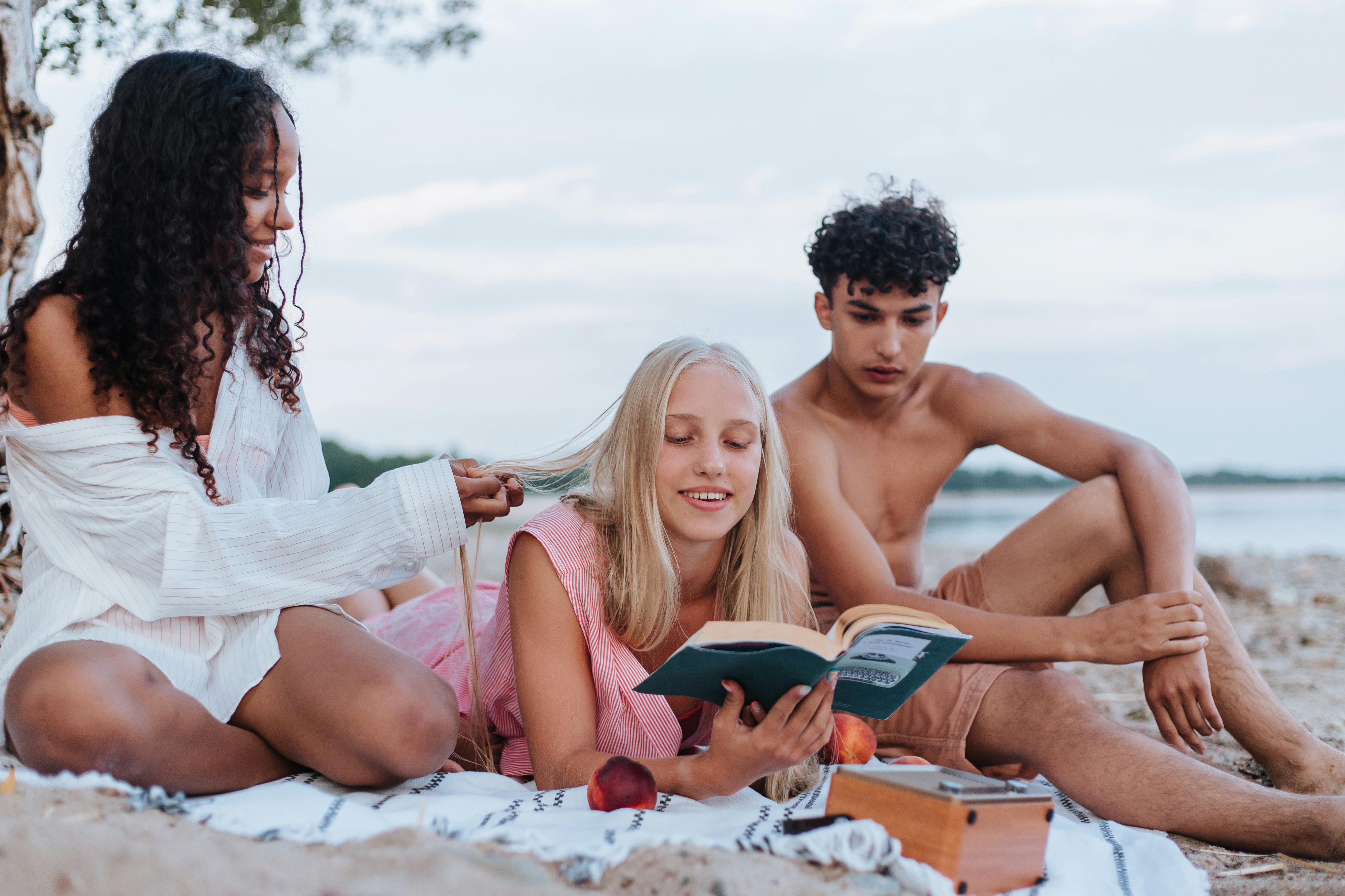 Three teenagers relaxing by the beach, reading and enjoying a summer picnic together.