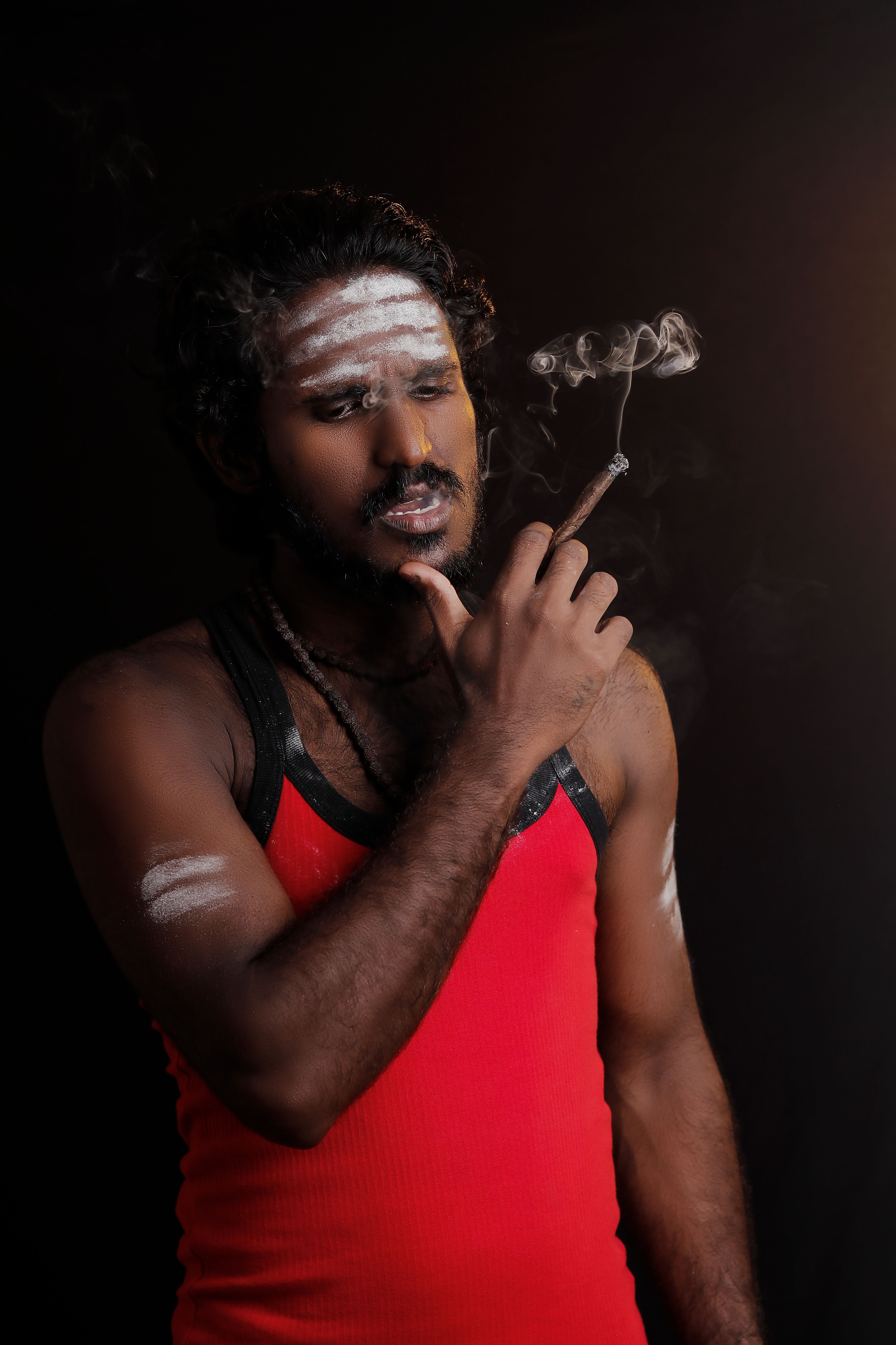 Moody portrait of a South Asian man with face paint, smoking indoors.