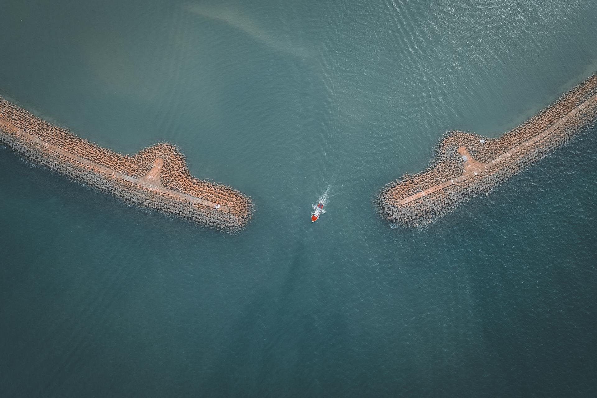 Aerial view of a lone boat navigating between two breakwaters in a calm bay.