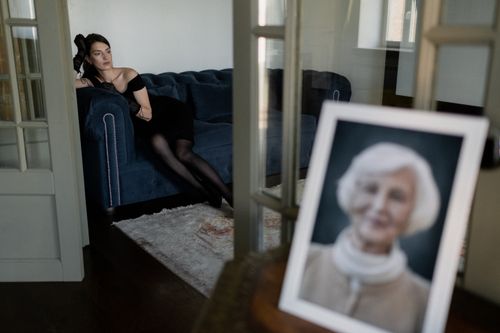 Free A woman in a black dress sits on a couch, gazing thoughtfully, with a framed portrait in the foreground. Stock Photo