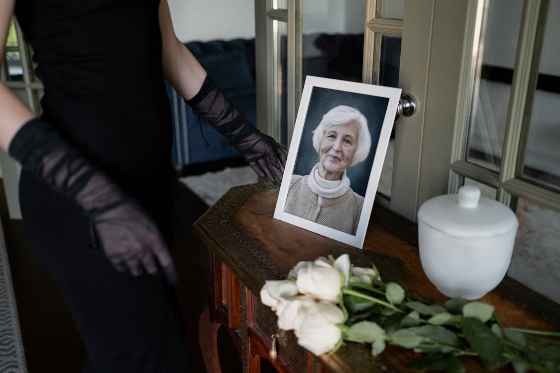 Free A reflective moment at a memorial with a framed portrait of an elderly woman, flowers, and an urn. Stock Photo