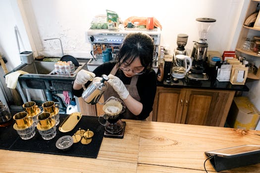 Asian barista wearing apron diligently pours drip coffee at café counter.