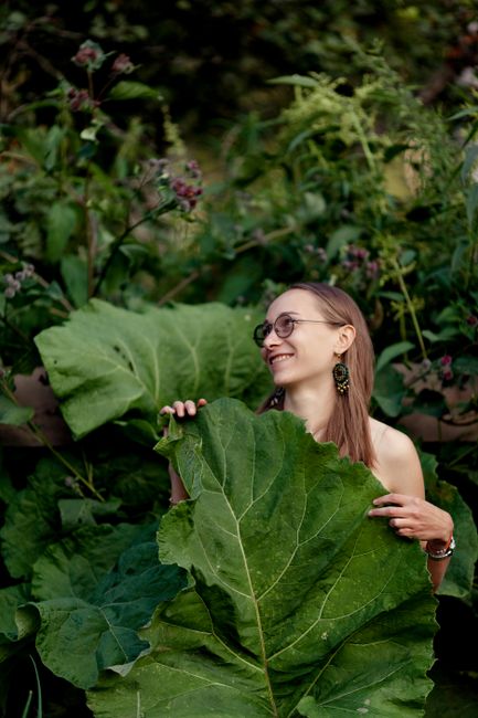 A woman smiling and holding a large green leaf outdoors, surrounded by lush greenery.
