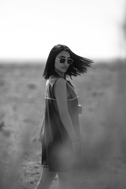 Monochrome image of a woman with sunglasses in a windswept dress looking back outdoors.