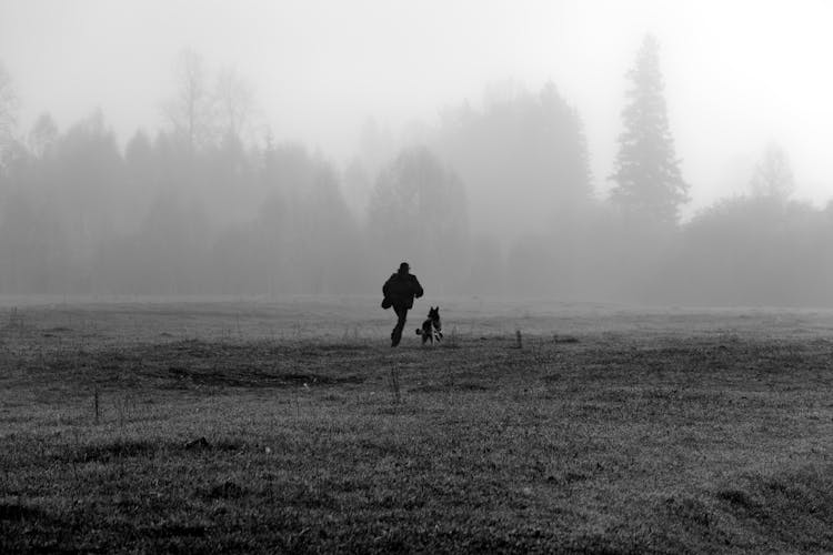 Grayscale Photo Of A Person Running With A Dog On Grassland