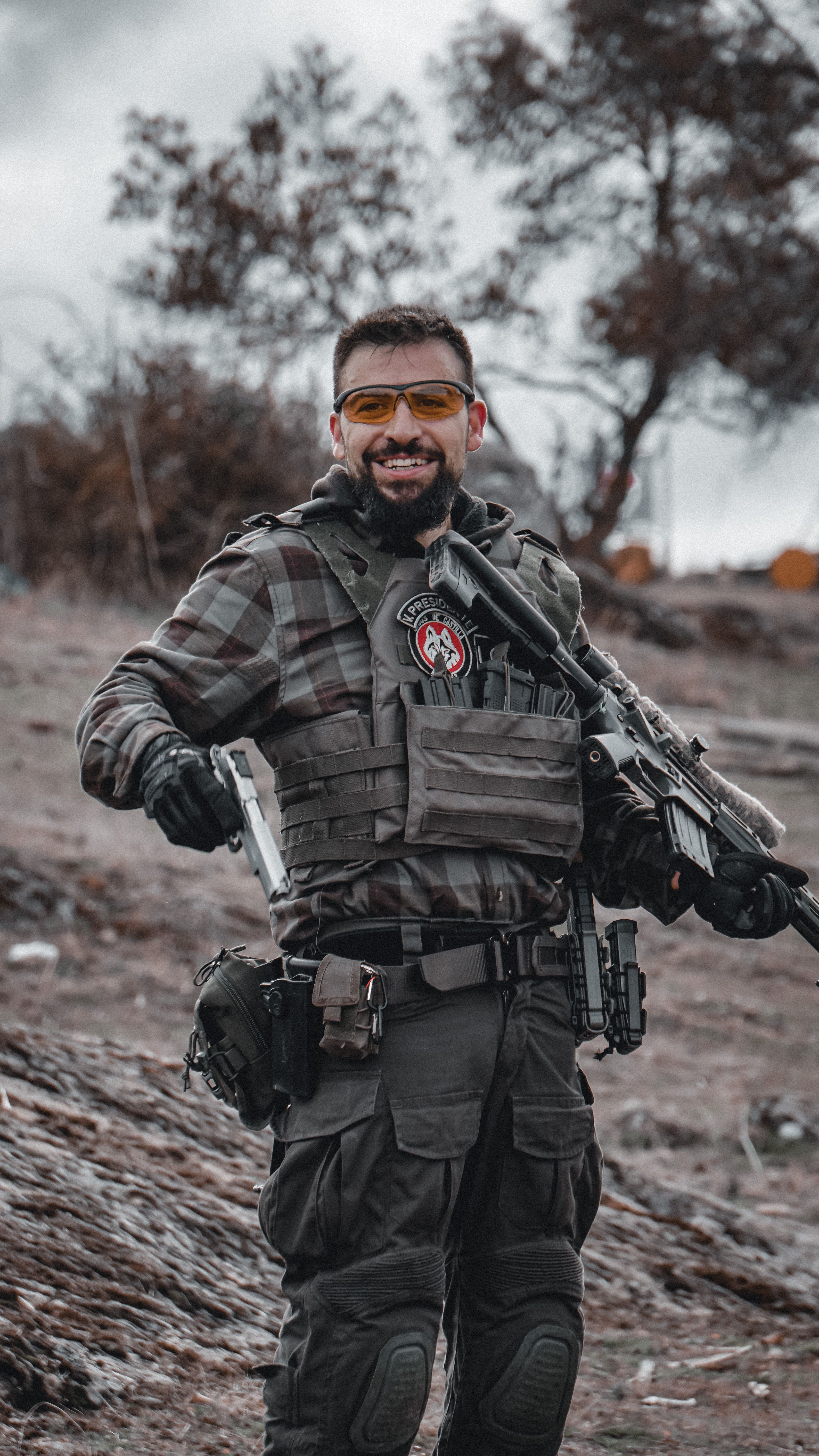 Smiling man in military uniform and tactical gear holding firearms outdoors.