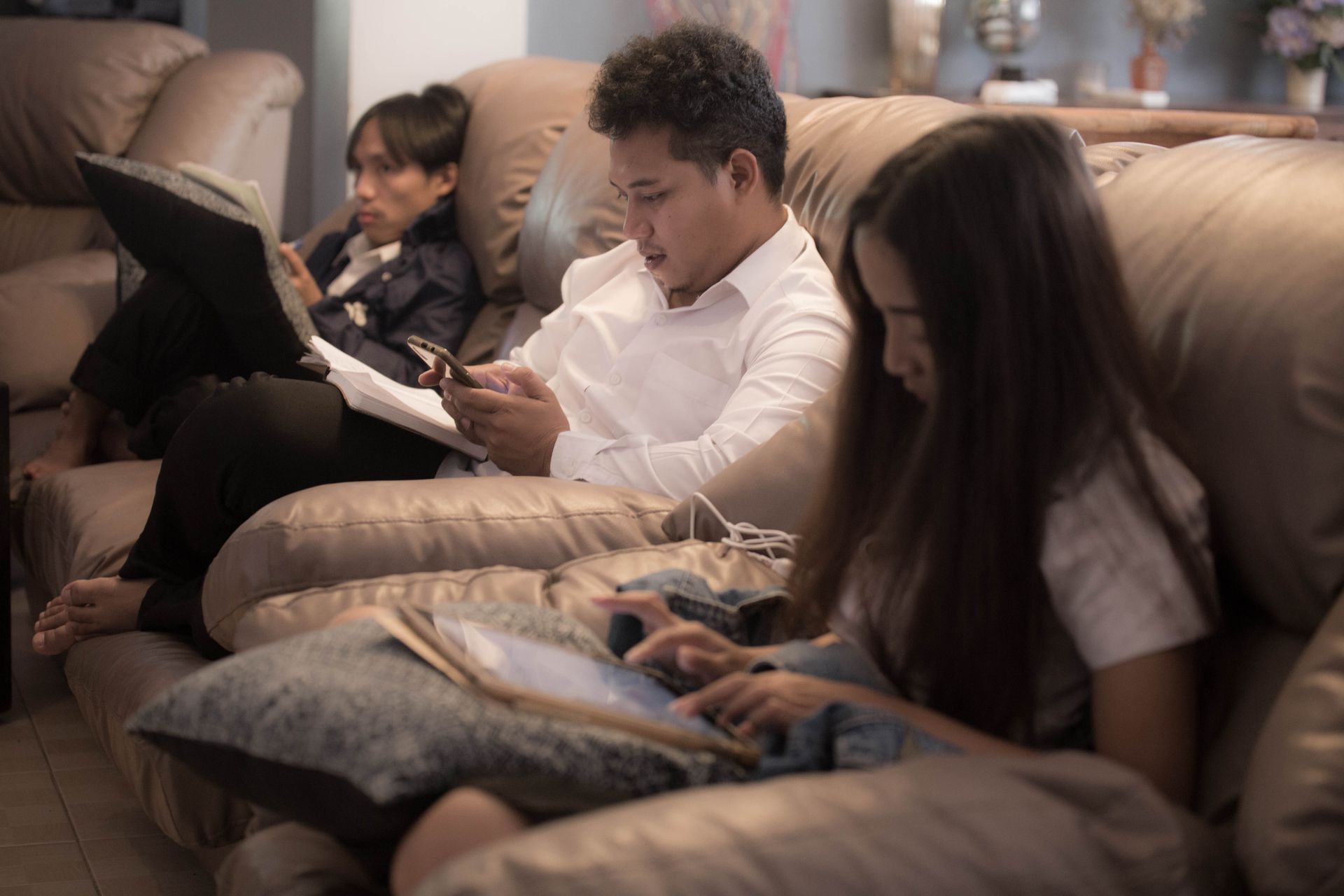 Three people relaxing indoors, using smartphones and reading on a sofa.